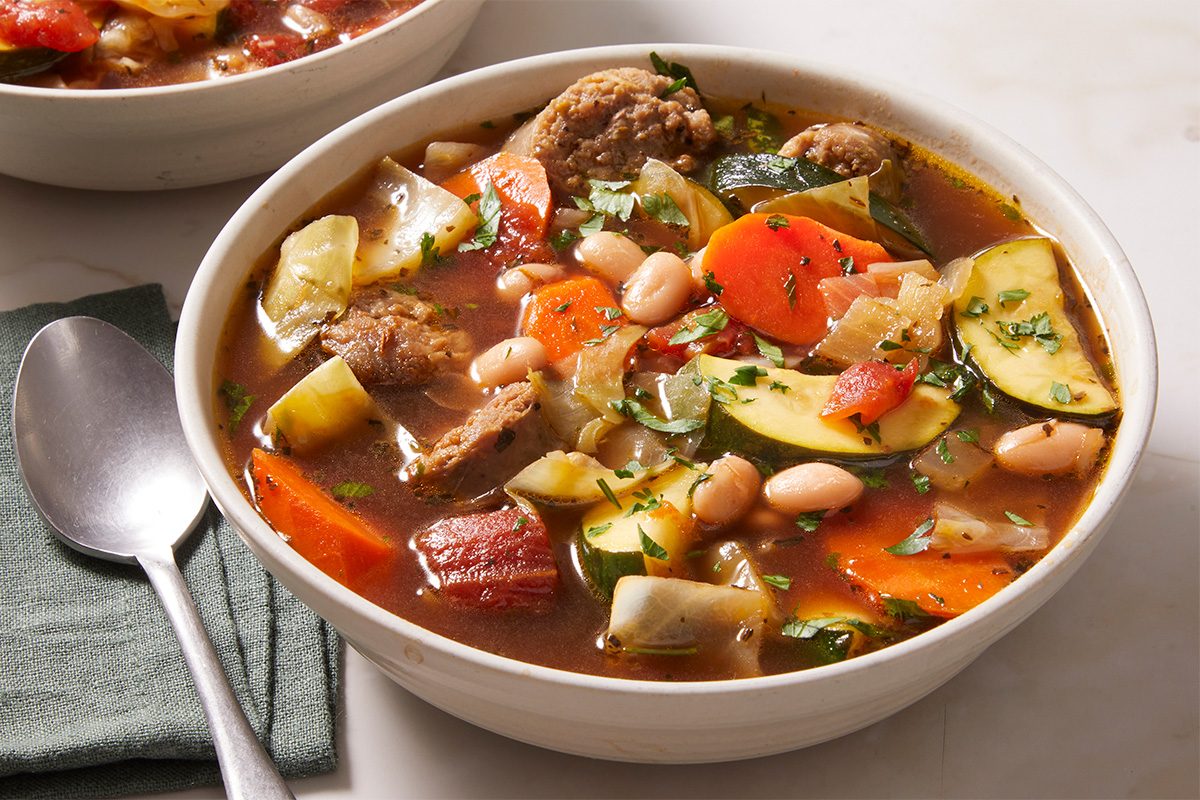 Close-up shot of a bowl of Minestrone Soup served hot, filled with vegetables and meat in a tomato-based broth, placed on a cloth napkin with a spoon alongside.