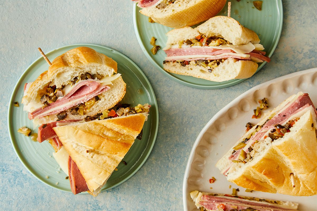 Overhead shot of mini muffuletta sandwiches on green plates and a white platter; The sandwiches are layered with deli meats, cheese, and olive salad filling, cut into wedges and secured with toothpicks;