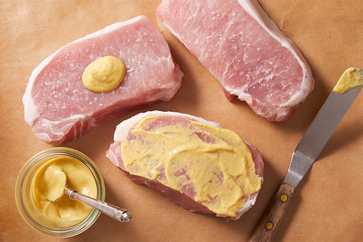 Overhead step-by-step food photography showing Dijon mustard being spread over raw pork loin chops during preparation for Dijon Pork Chops.