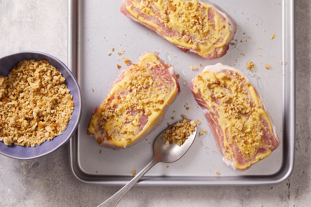 Overhead step-by-step food photography showing seasoned bread crumbs being added to mustard-coated pork chops on a baking sheet.