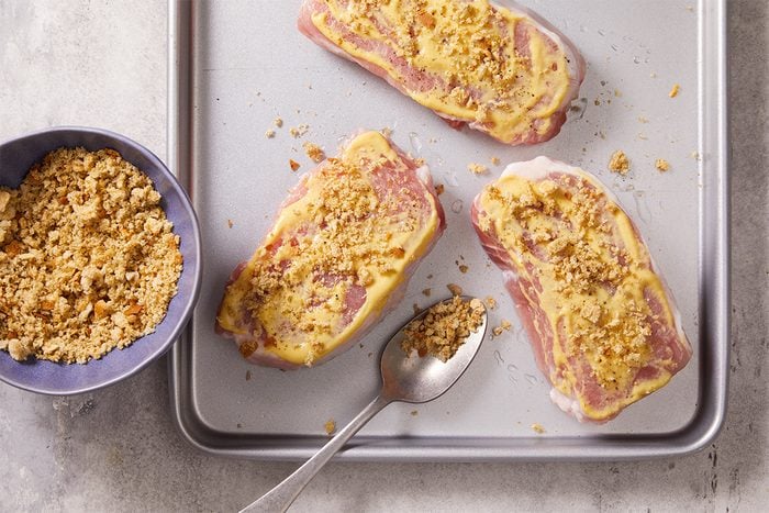 Overhead step-by-step food photography showing seasoned bread crumbs being added to mustard-coated pork chops on a baking sheet.