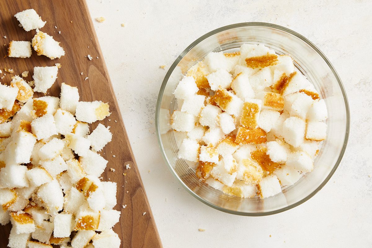 A glass bowl filled with cubes of white bread, some with crusts, sits on a light surface next to a cutting board with more cubed bread scattered on it.