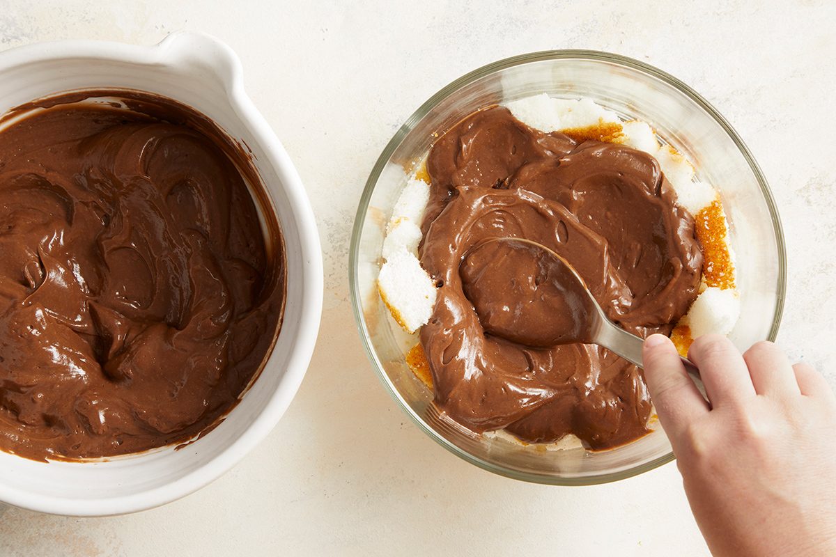 A hand spreads chocolate pudding over a layer of cake cubes in a glass bowl, with an additional bowl of chocolate pudding nearby on a light surface.