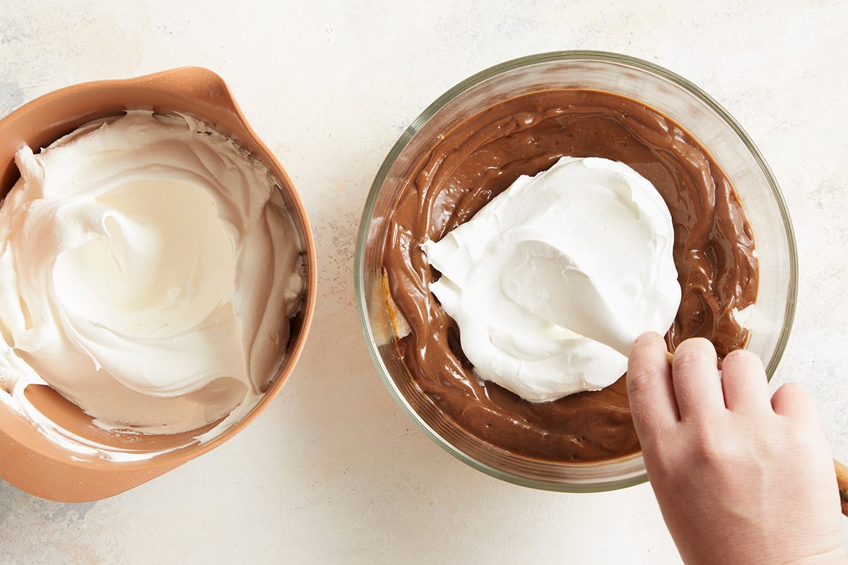 A hand folds whipped cream into a bowl of chocolate mixture, with a separate bowl of whipped cream beside it on a light-colored surface.