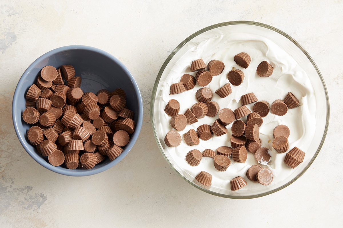 A bowl of whipped cream topped with mini peanut butter cups sits next to a separate bowl filled with more mini peanut butter cups on a light surface.