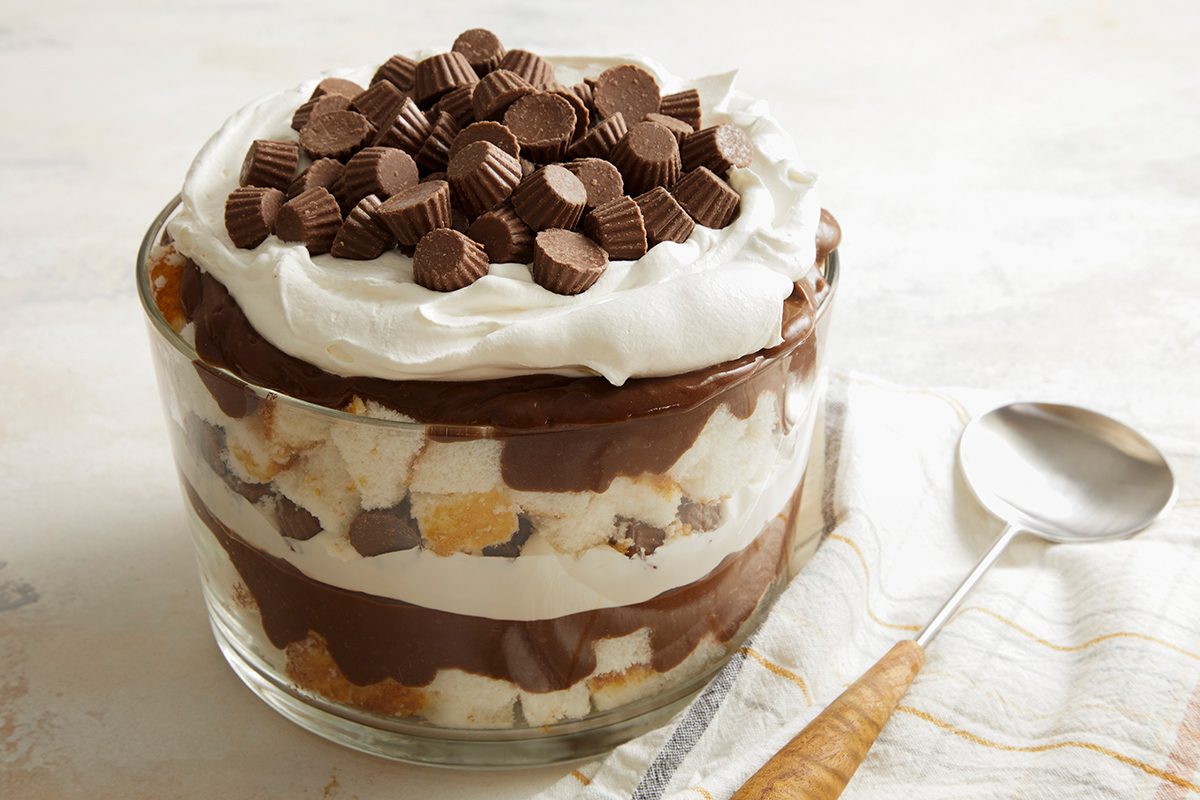 A glass trifle dish filled with layered chocolate pudding, whipped cream, cake pieces, and topped with mini peanut butter cups sits next to a metal spoon on a white striped towel.