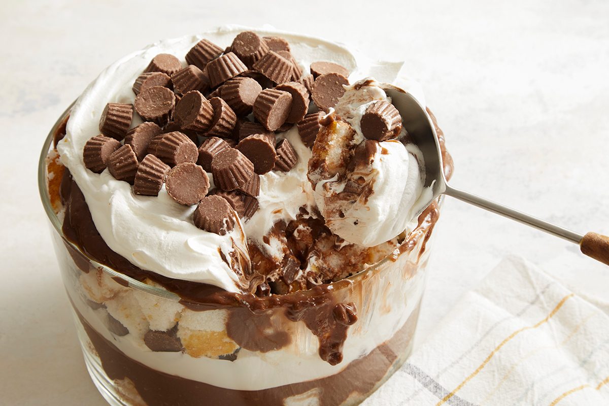 A glass bowl filled with layered chocolate pudding, whipped cream, cake pieces, and topped with mini peanut butter cups. A spoon is scooping out a portion of the dessert.