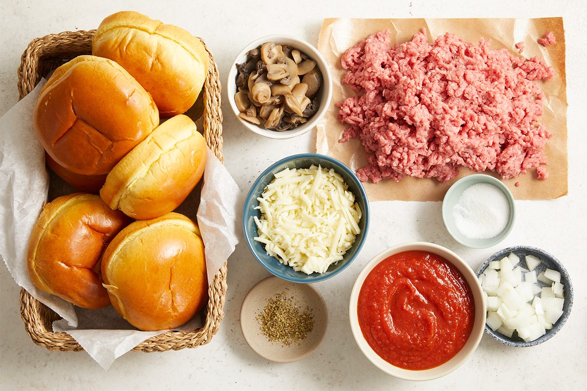 Overhead, horizontal image showing all ingredients arranged on a light surface for Open-Faced Pizza Burgers. Ingredients include raw ground beef, chopped onion, pizza sauce, drained mushroom stems and pieces, shredded mozzarella cheese, seasonings, and split hamburger buns. This flat lay establishes the full ingredient lineup before cooking begins and supports a fast-track, step-by-step recipe process.