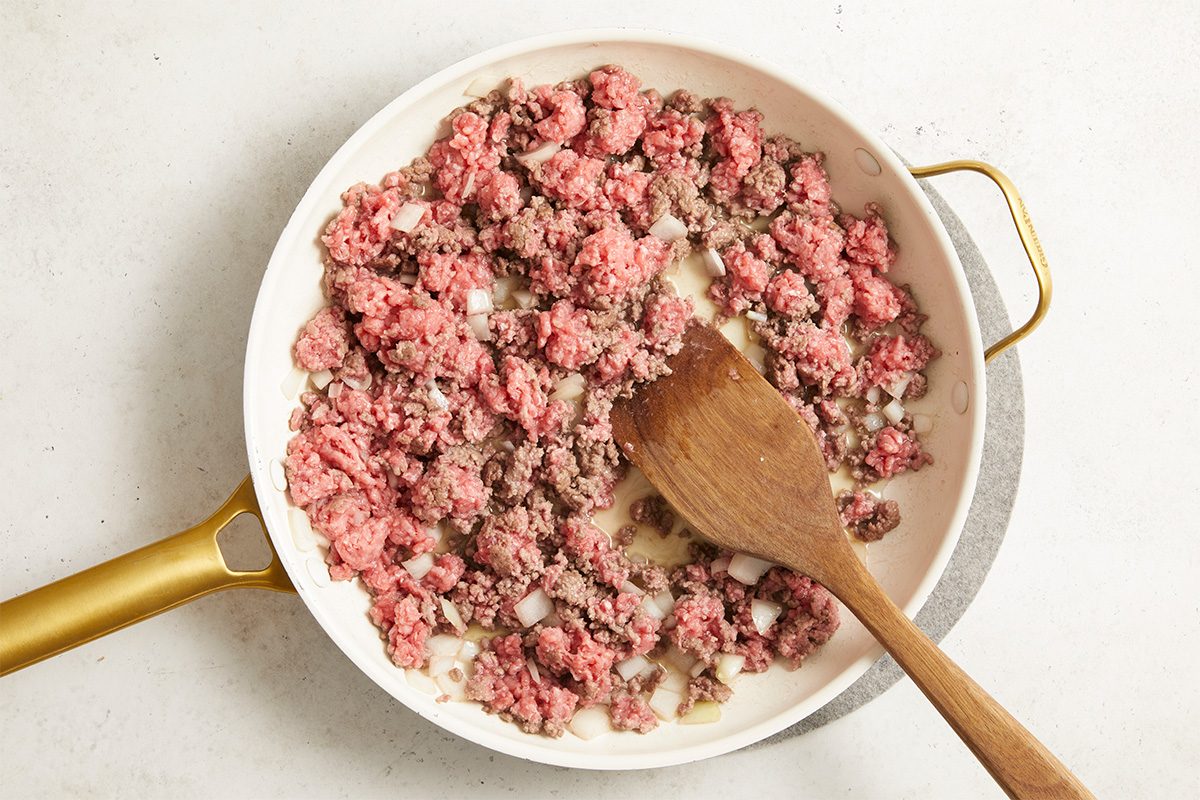 Overhead, horizontal step-by-step image of ground beef cooking in a skillet. A wooden spoon stirs the beef as it begins to brown, marking the first active cooking step in preparing Open-Faced Pizza Burgers. The image emphasizes texture and doneness during the fast-track cooking process.