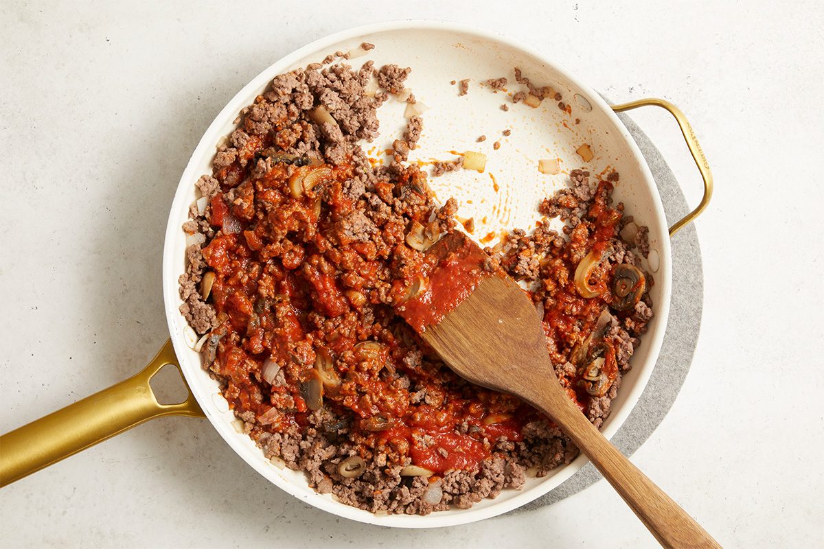 Overhead, horizontal image showing browned ground beef in a skillet as pizza sauce and seasonings are added. The mixture is partially combined with visible sauce and spices, illustrating the transition from plain beef to a seasoned pizza-style filling for Open-Faced Pizza Burgers.