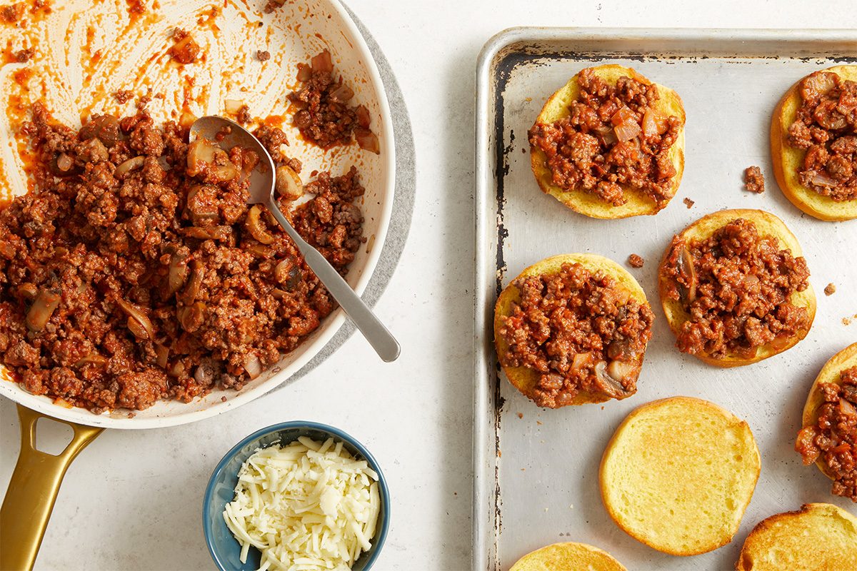 Overhead, horizontal step-by-step image showing finished pizza beef mixture alongside toasted hamburger buns on a baking sheet. The composition highlights preparation for assembly, with the cooked filling ready to be spooned onto the buns for Open-Faced Pizza Burgers.