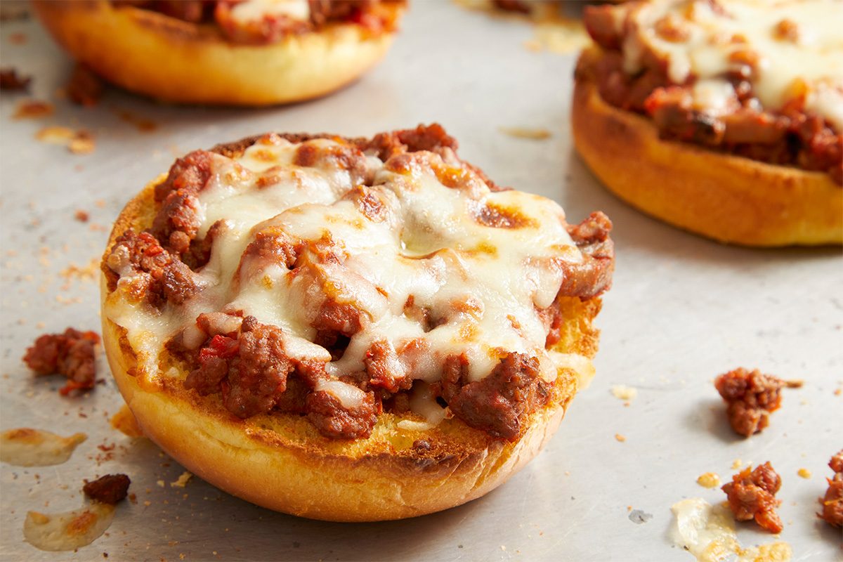 Overhead, horizontal hero-style close-up of a finished open-faced pizza burger on a baking sheet. Melted mozzarella cheese tops the savory pizza beef mixture on a toasted bun, emphasizing texture, color, and final presentation.