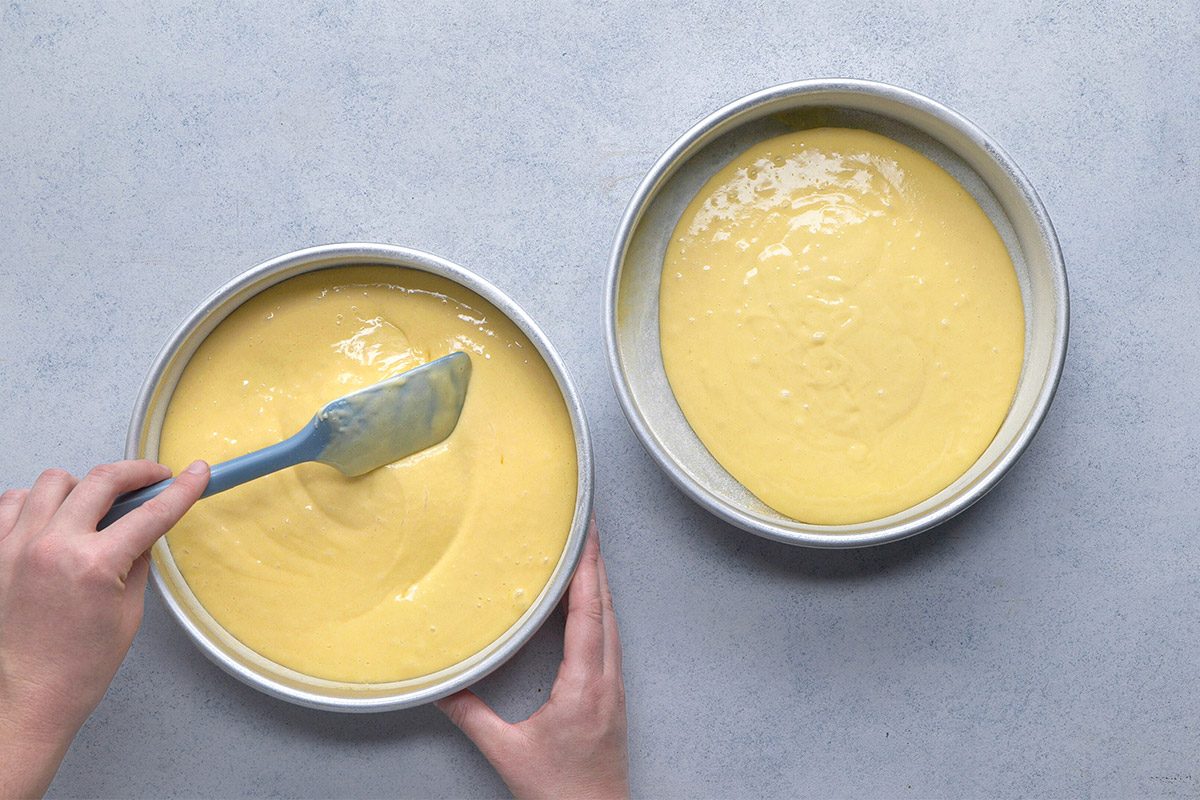 Overhead shot of two round cake pans filled with batter; a person smooths the batter in one pan with a blue spatula while steadying the pan with their other hand;