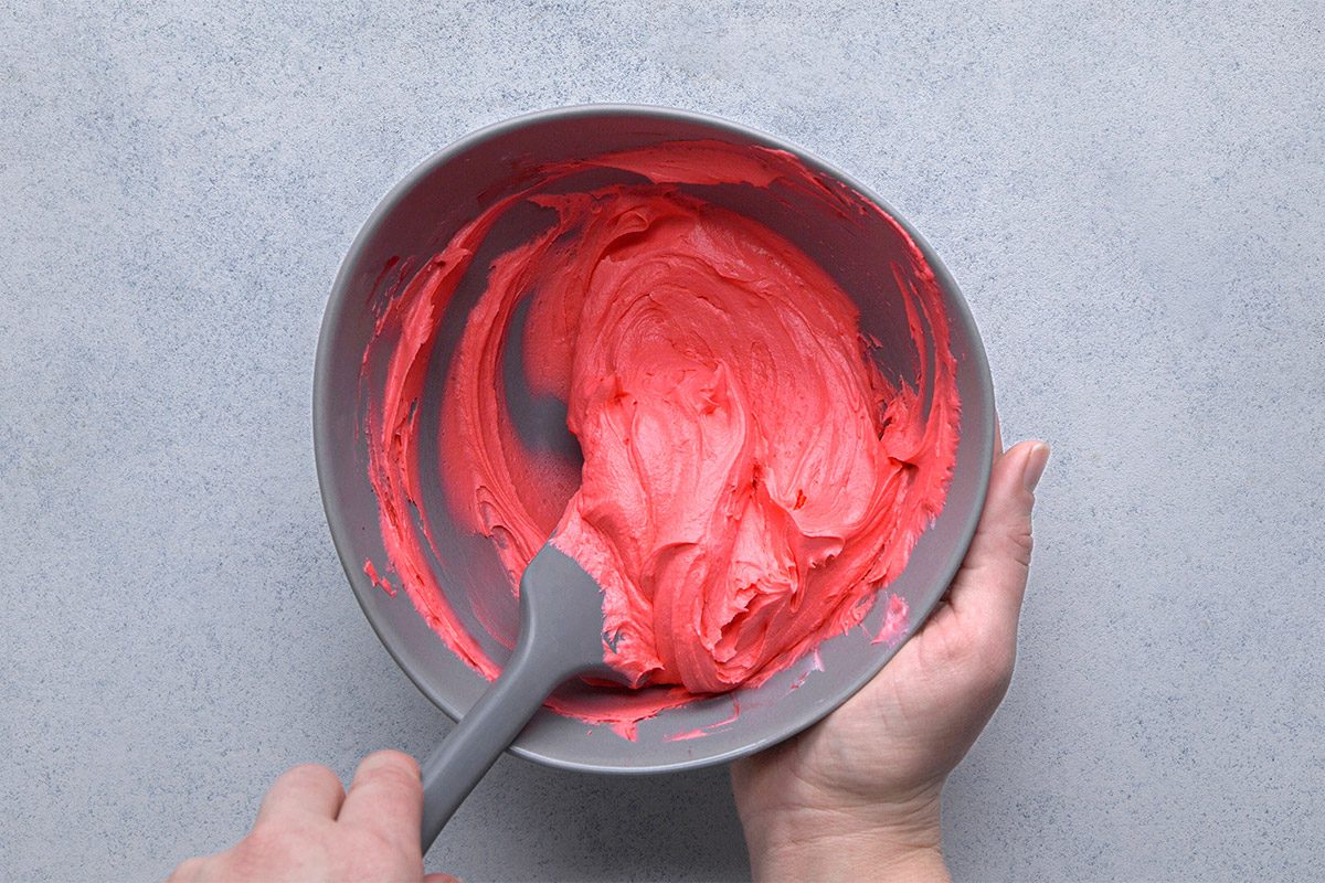 Overhead shot of a person holding a bowl of bright pink frosting or batter, mixing it with a spatula over a light-colored surface;