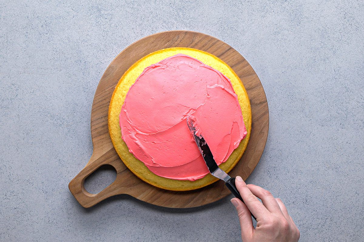 Overhead shot of a hand spreading pink frosting on a round yellow cake set on a wooden cutting board over a light-colored surface;