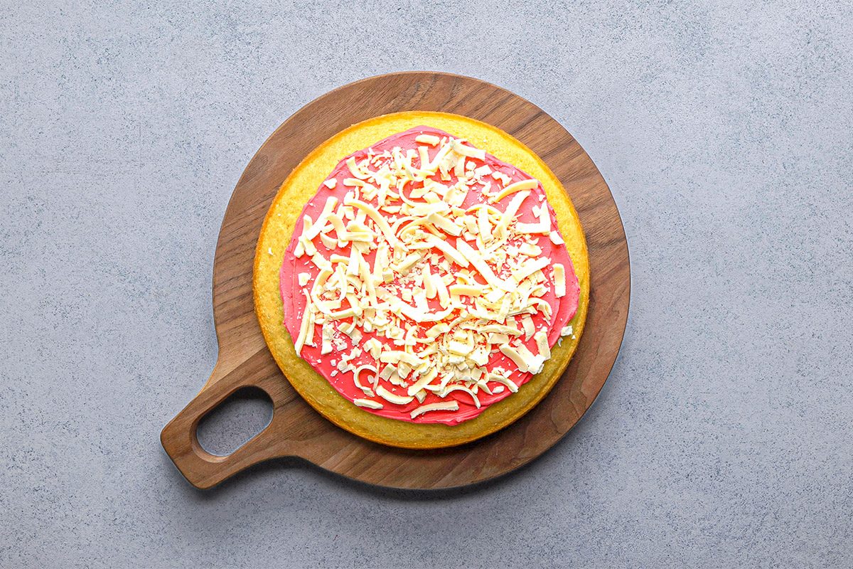 Overhead shot of a round cake with pink frosting and white chocolate shavings on a wooden serving board against a light background;