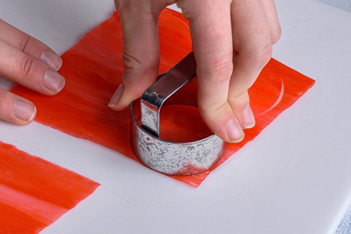 Close shot of a person pressing a round metal cookie cutter into a sheet of bright red material on a white surface, preparing to cut out a circular shape;