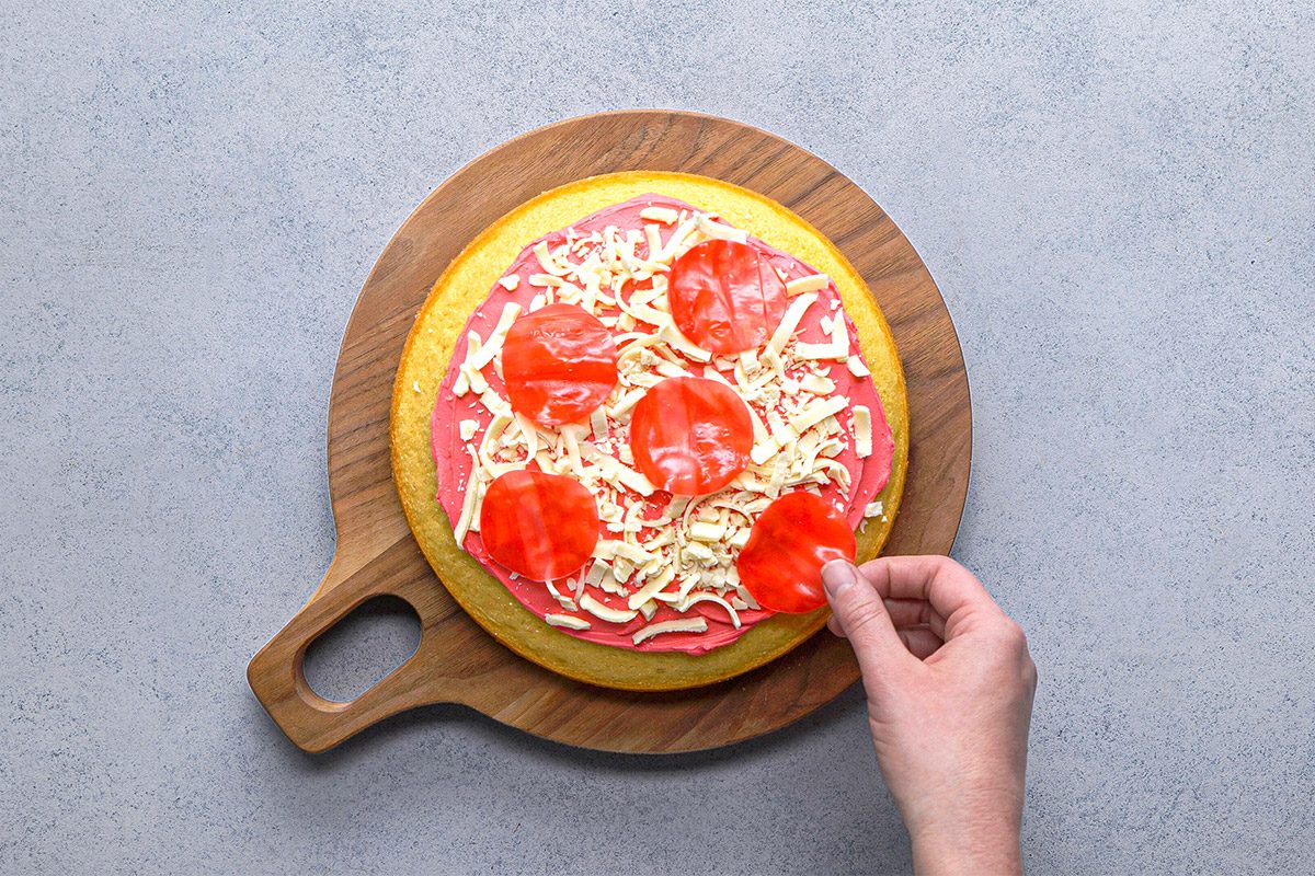 Overhead shot of a hand placing a slice of pepperoni onto an uncooked pizza topped with shredded cheese and tomato sauce on a round wooden board;