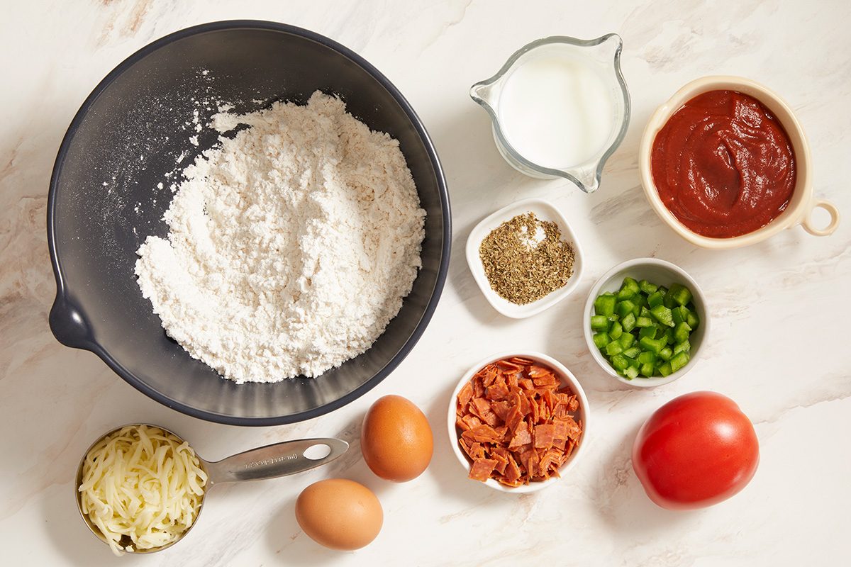 A bowl of flour, a small pitcher of milk, a bowl of tomato sauce, shredded cheese, dried herbs, chopped green peppers, chopped pepperoni, two eggs, and a whole tomato arranged on a white surface.