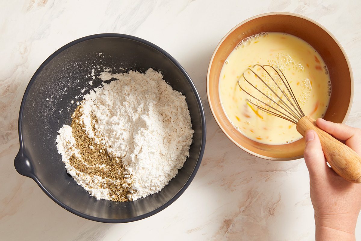 A hand whisking eggs in a bowl next to a black bowl containing flour and herbs on a light-colored countertop.