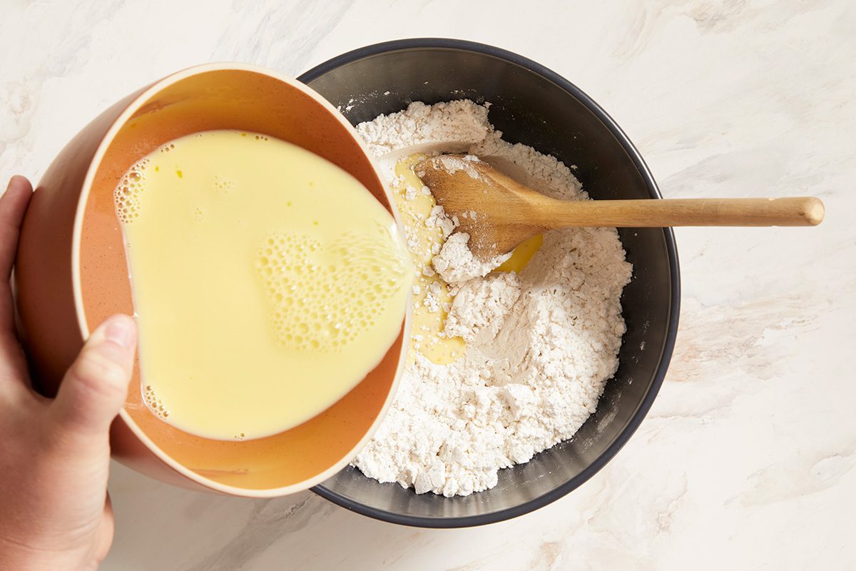 A hand pours a bowl of beaten eggs into a large bowl of flour, which also contains a wooden spoon, on a light countertop.