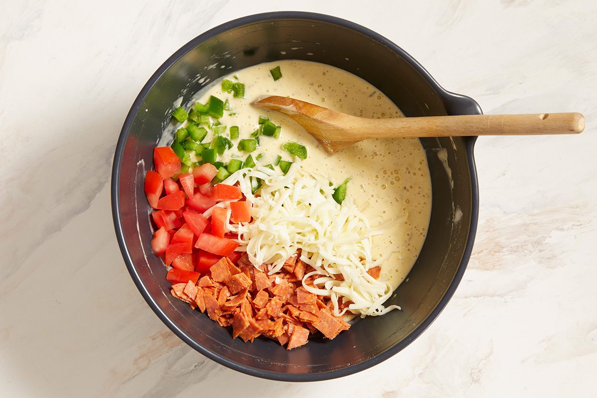 A black mixing bowl contains a creamy batter topped with chopped green peppers, diced tomatoes, shredded cheese, and diced cooked sausage. A wooden spoon rests on the bowl's edge.