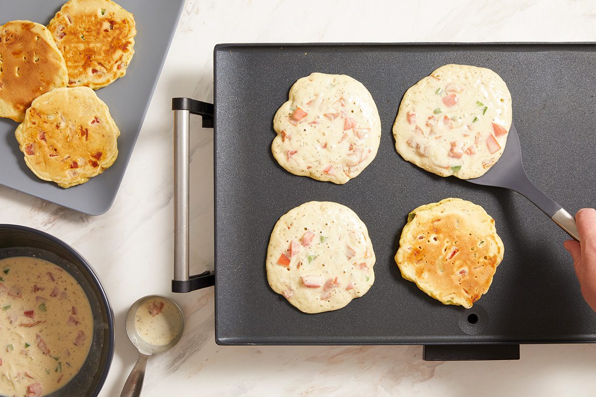 Four pancakes with visible bits of vegetables cook on a griddle, with one being flipped by a spatula. Nearby, more cooked pancakes rest on a plate, and a bowl of pancake batter sits beside them.