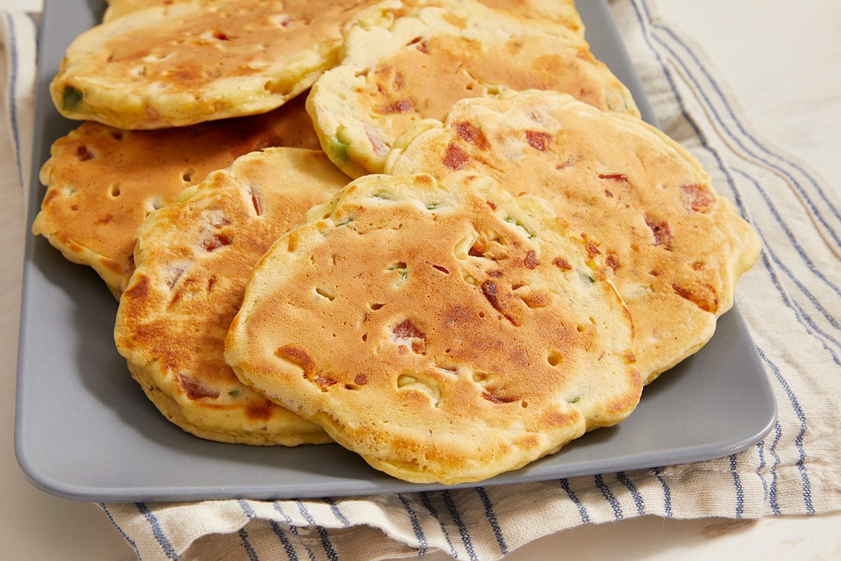 A gray plate holds several golden-brown savory pancakes with visible bits of ham and green vegetables, resting on a striped cloth napkin.
