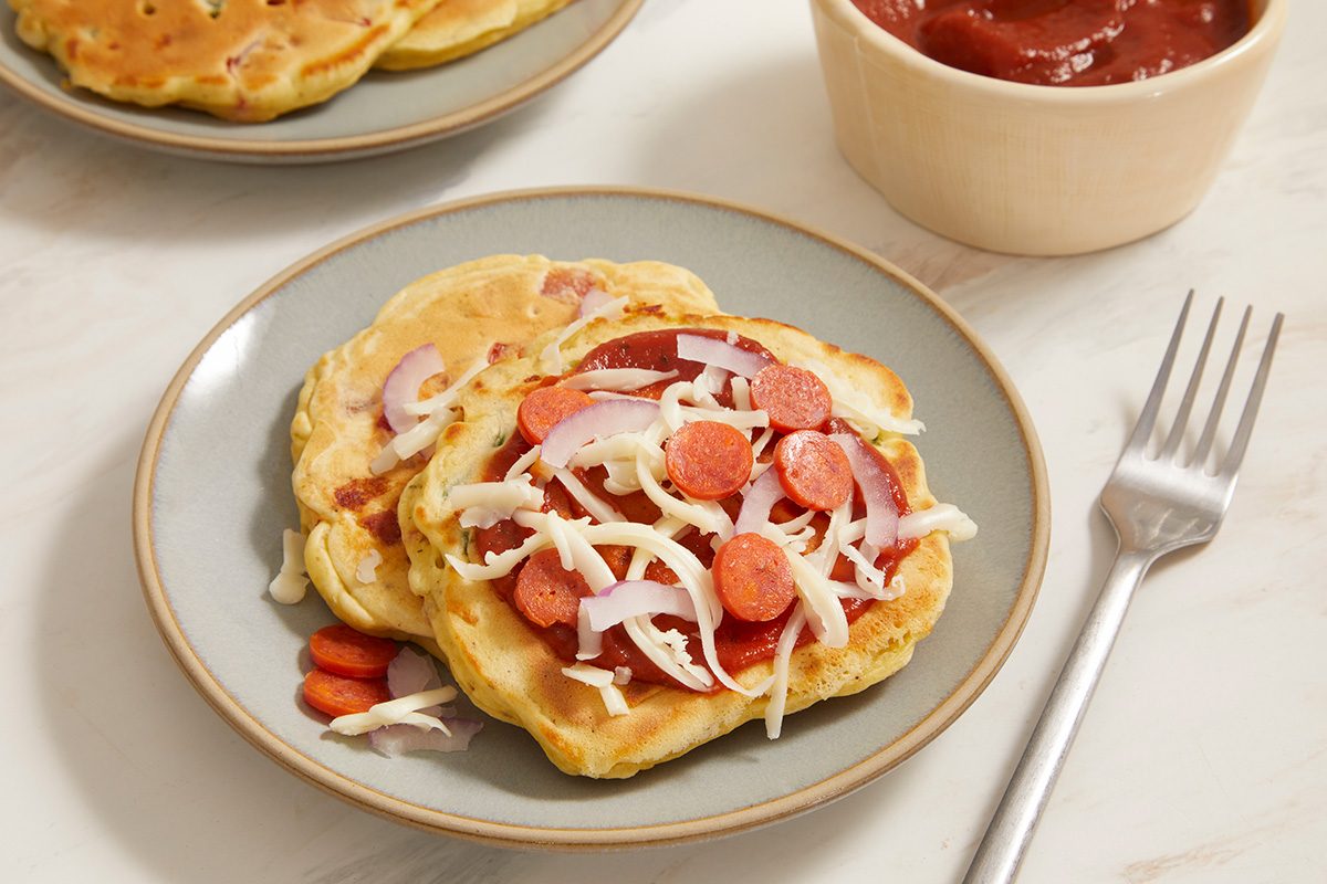 Two savory pancakes topped with sliced sausages, shredded cheese, and thinly sliced red onions are served on a plate with a fork nearby. A bowl of red sauce is in the background.