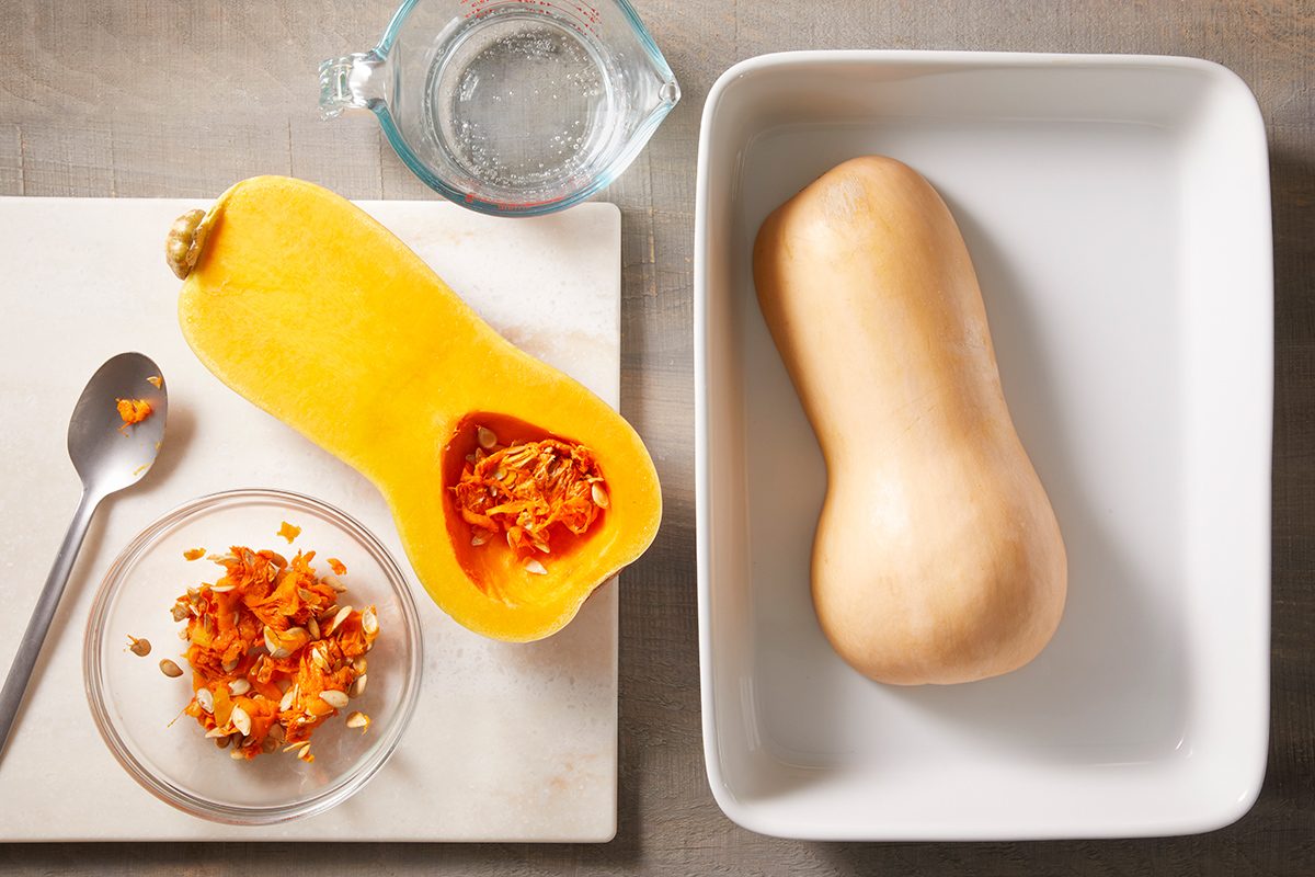 A whole butternut squash and a halved butternut squash on separate surfaces, with seeds and pulp in a glass bowl, a spoon, and a measuring cup of water nearby.