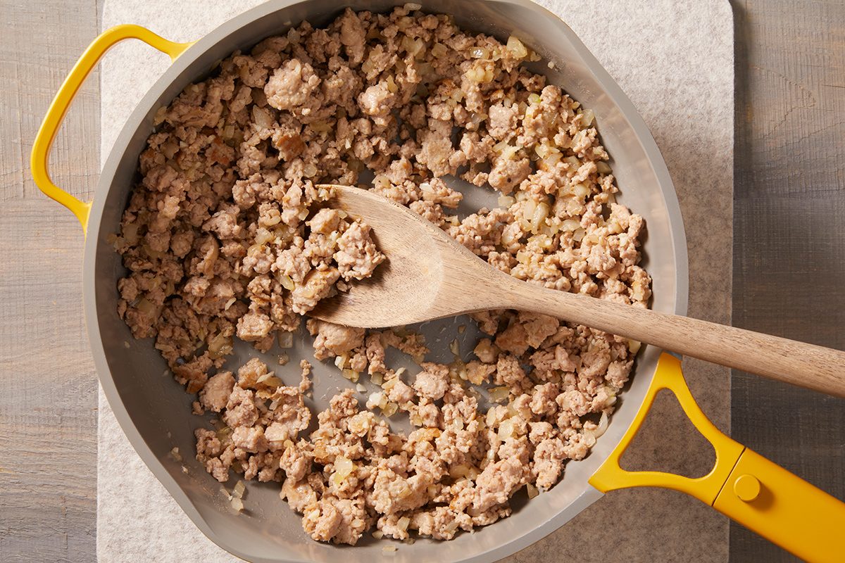Cooked ground turkey in a gray skillet with yellow handles, being stirred with a wooden spoon. The skillet is placed on a light-colored surface.