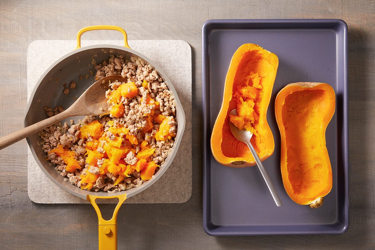 A yellow pot with cooked rice and chunks of squash is next to a baking tray holding two halved, roasted butternut squash, one with some flesh scooped out and a spoon resting inside.