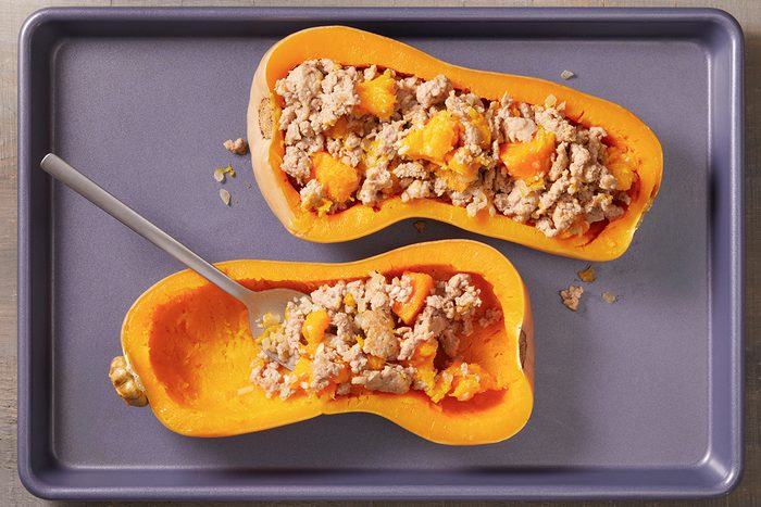 Two halves of butternut squash filled with a ground meat mixture are placed on a baking sheet. One half has a spoon in it, showing some of the filling scooped out.