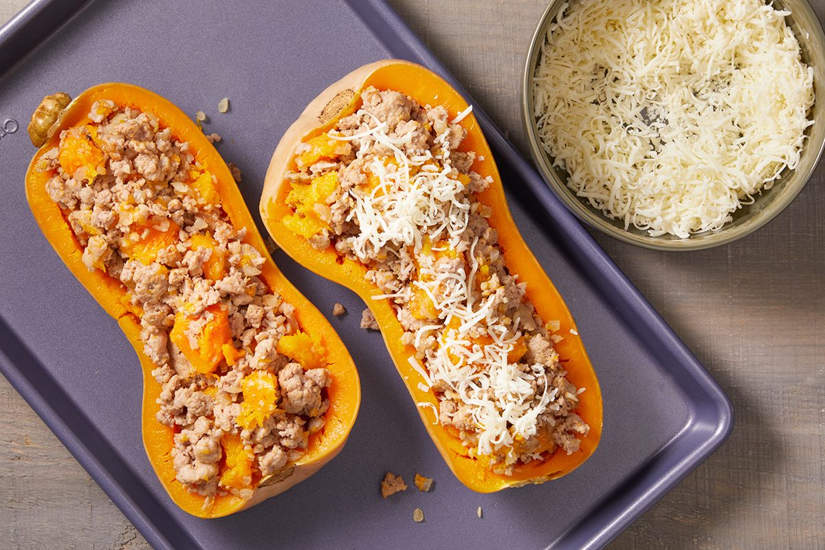 Two halves of butternut squash stuffed with a mixture of ground meat and topped with shredded cheese are placed on a baking tray. A bowl of shredded cheese sits nearby.