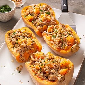 Four halved butternut squashes stuffed with a mixture of rice, ground meat, cheese, and herbs are arranged on a white platter, garnished with chopped parsley. A serving spatula and small bowls are nearby.