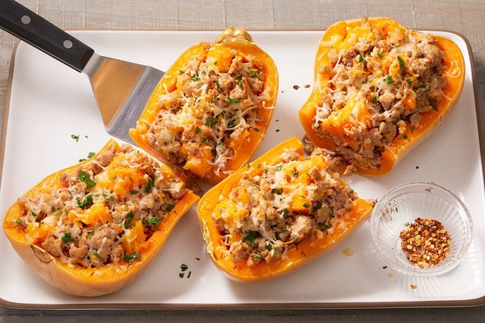 Four baked butternut squash halves on a white tray, stuffed with a mixture of rice, vegetables, and melted cheese. A metal spatula rests beside them, and a small dish of red pepper flakes is nearby.