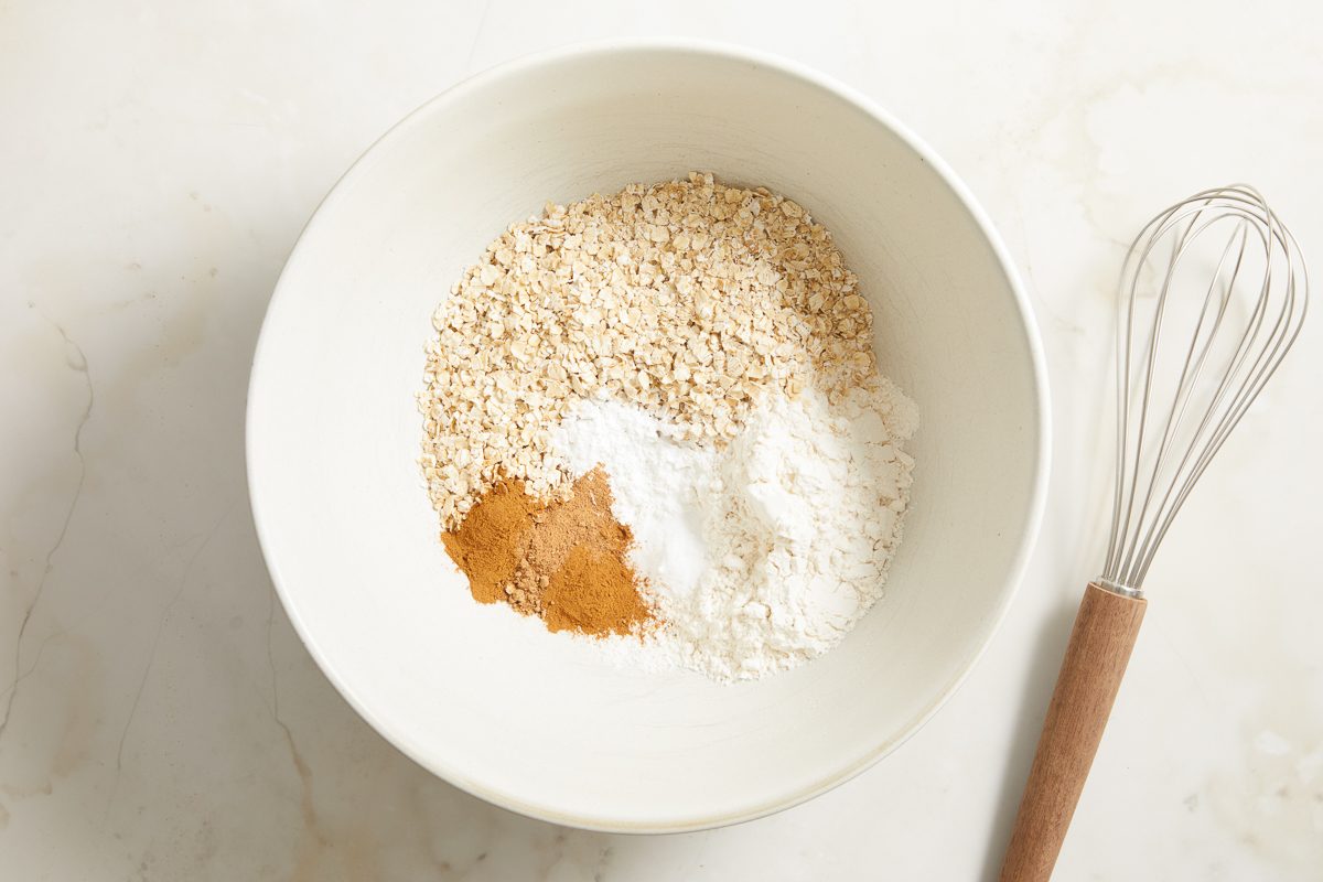 flour, oats, baking powder, baking soda, cinnamon and nutmeg in a large bowl next to a whisker