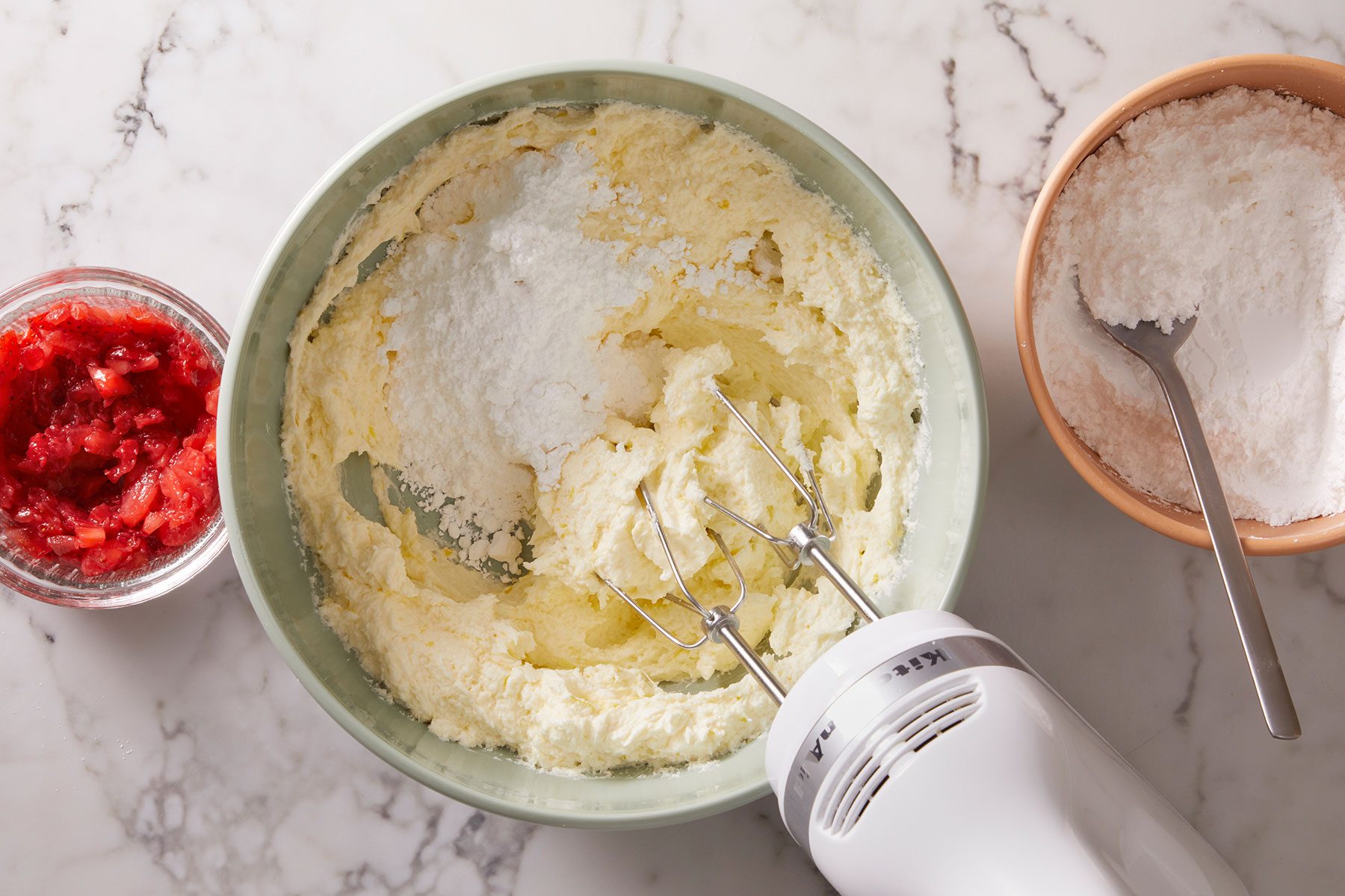 Beating cream, sugar, butter and lemon zest in a bowl with some chopped strawberries and sugar placed on side.