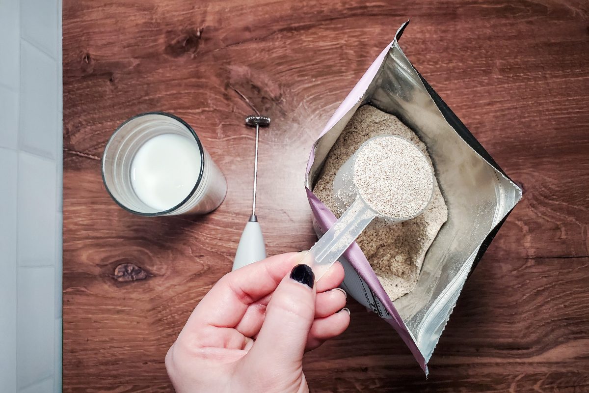 A hand holds a scoop of protein powder above an open bag, next to a glass of milk and a small electric whisk, all placed on a wooden surface.