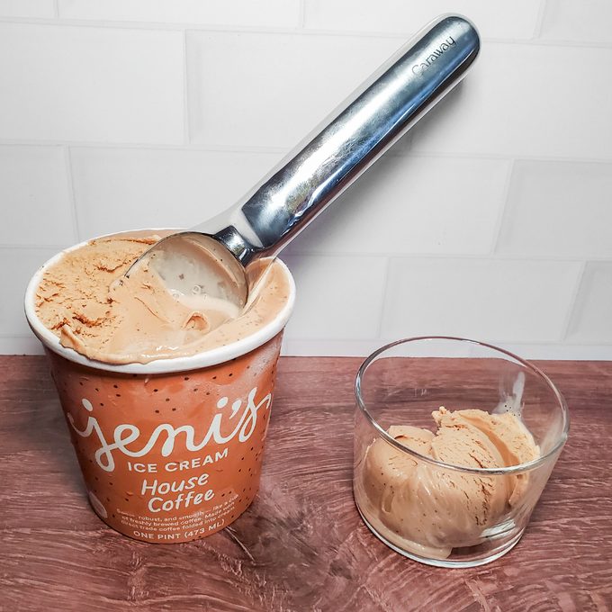 A container of Jeni’s House Coffee ice cream with a scoop inside is next to a small glass bowl holding a scoop of the ice cream, all on a wooden surface with a white tiled background.
