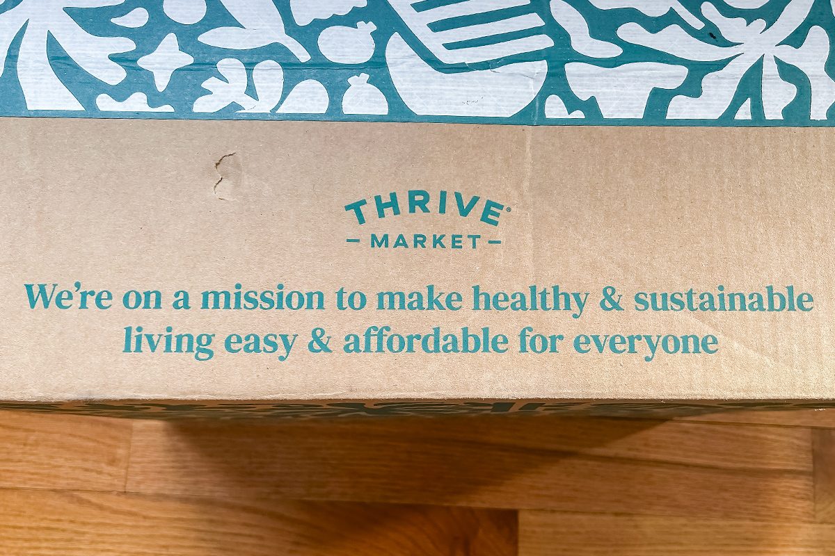 A cardboard Thrive Market box with teal text that reads: "We're on a mission to make healthy & sustainable living easy & affordable for everyone." The box rests on a wooden floor.