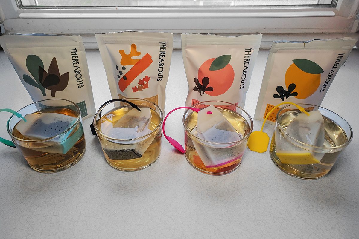 Four glass cups of tea with different colored tea bag labels sit in front of matching tea packages illustrated with leaves and fruits, displayed on a white surface by a window.