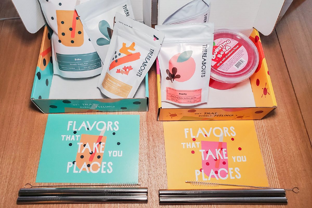 Two colorful food boxes with snacks, chopsticks, and postcards that read "FLAVORS THAT TAKE YOU PLACES" are displayed on a wooden table. The boxes and packaging feature playful fruit designs and bright colors.