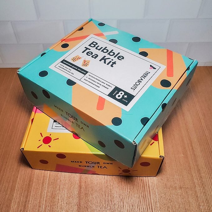 Two colorful boxes on a wooden surface. The top box is teal with black dots, labeled "Bubble Tea Kit" for ages 18+, while the bottom box is yellow with a red sun design.