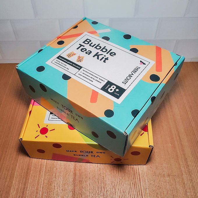 Two colorful boxes on a wooden surface. The top box is teal with black dots, labeled "Bubble Tea Kit" for ages 18+, while the bottom box is yellow with a red sun design.