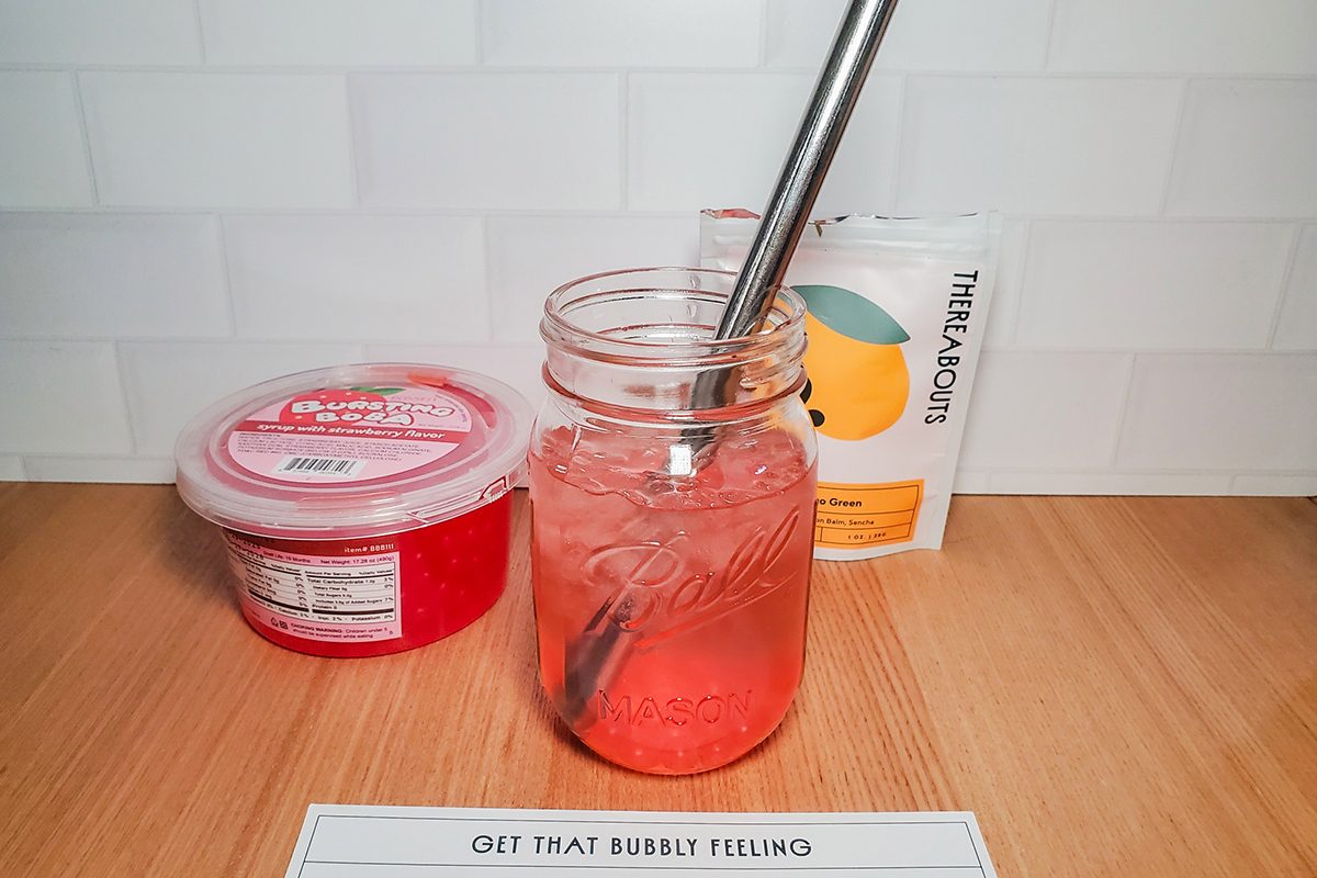 A mason jar filled with pink fizzy drink and a metal straw sits on a wooden counter. Behind it are a container of red gelatin dessert and a packet labeled “TRUE LEMON.” A label reads, “GET THAT BUBBLY FEELING.”.