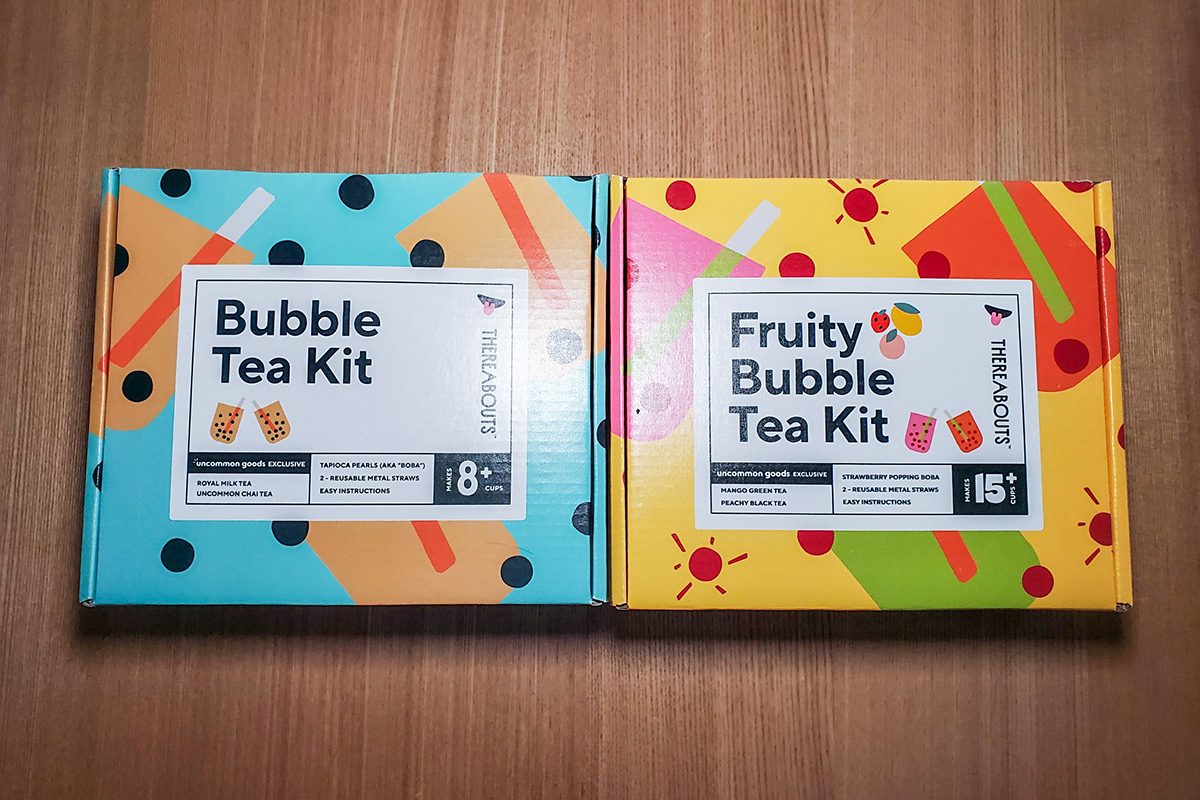 Two colorful boxes on a wooden surface. The left box is blue with black dots and orange shapes, labeled "Bubble Tea Kit." The right box is yellow with red dots and suns, labeled "Fruity Bubble Tea Kit.
