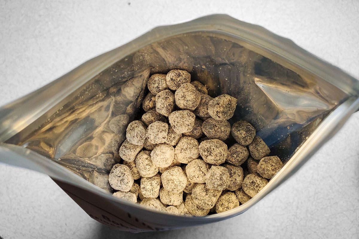 An open resealable bag filled with round, brown, crumbly treats or pellets, viewed from above on a light-colored surface.