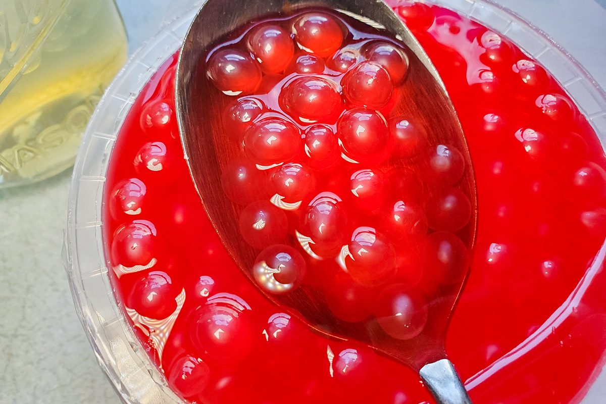 A close-up of a spoon scooping shiny, red maraschino cherries in thick syrup from a glass bowl. A portion of a yellow drink in a glass is visible in the background.