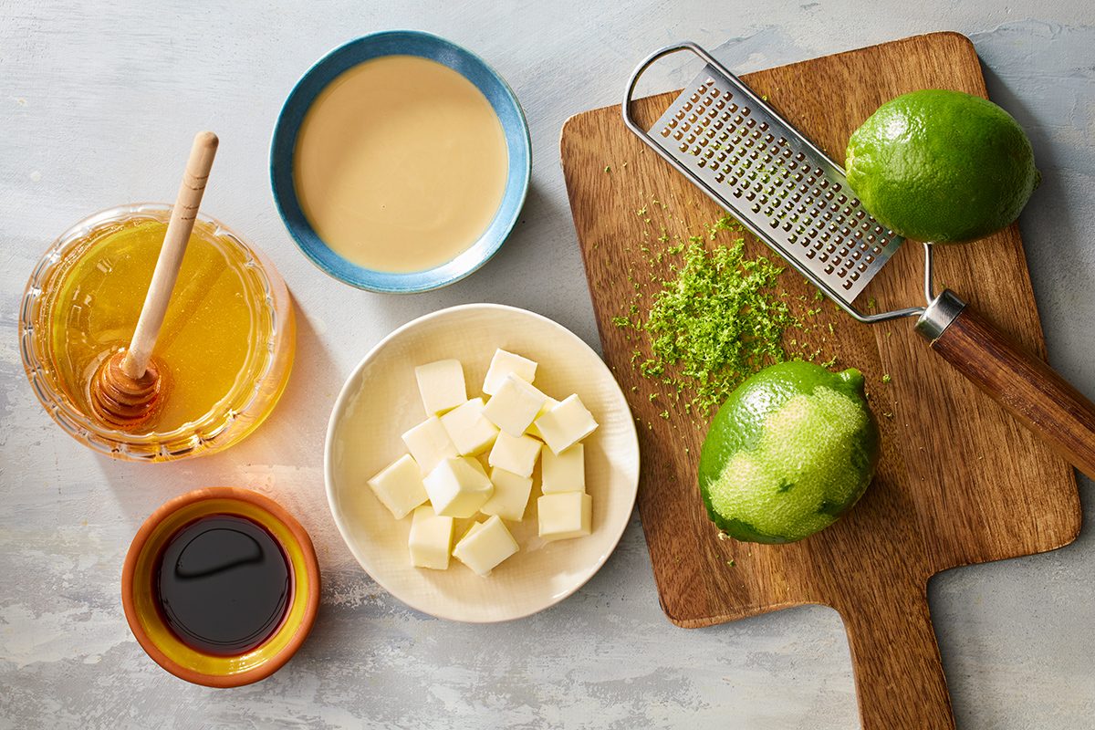 A cutting board with grated lime zest, whole limes, and a grater is surrounded by bowls of honey, cubed butter, tahini, and a dark liquid, all set on a light surface.